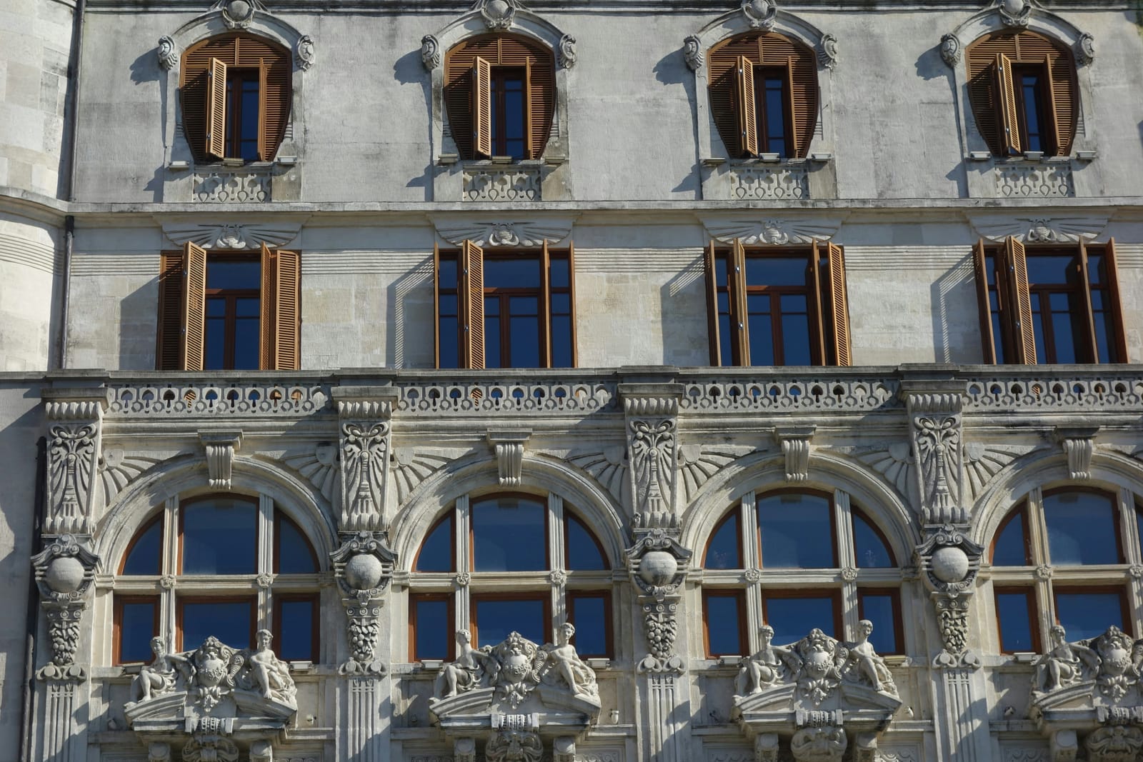 Gazdagon tagolt historizáló homlokzat közelről: íves ablakkeretezések, konzolos párkányok, plasztikus díszítőelemek és faspaletták – az a felületgeometria, amelyre a táblás hőszigetelő rendszerek geometriai és örökségvédelmi okokból nem vihetők fel