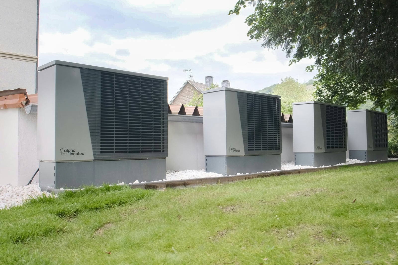 Four high-capacity air-to-water heat pump outdoor units in cascade beside an existing rendered building – design heat output follows from the building's heat loss, so an unchanged envelope shows up directly in the installed plant capacity